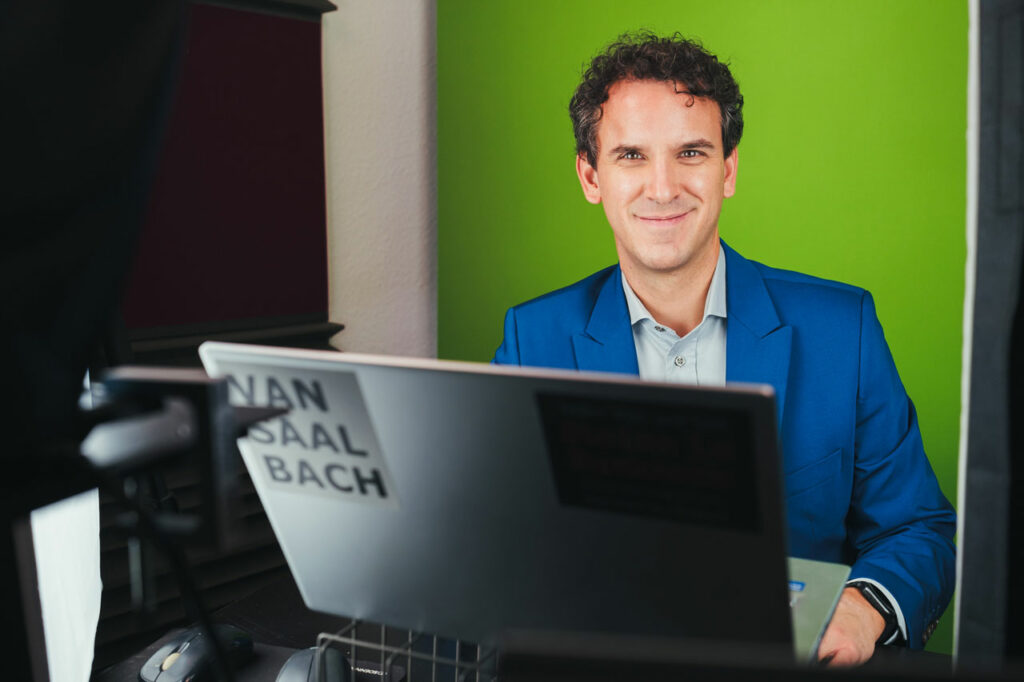 Ferry van Saalbach is sitting in a suit in front of a laptop, with a green screen in the background.